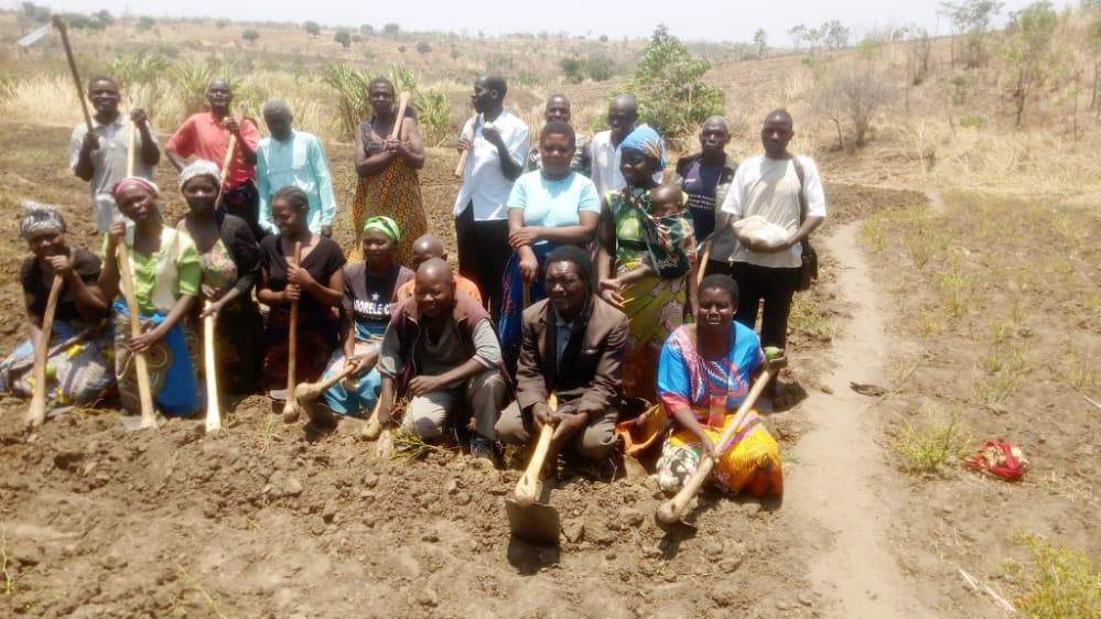 A group of person with farming equipment such as hoes on the field they are preparing for sowing. They are arranged in three rows with the foremost row kneeling and the person in the other two rows standing.