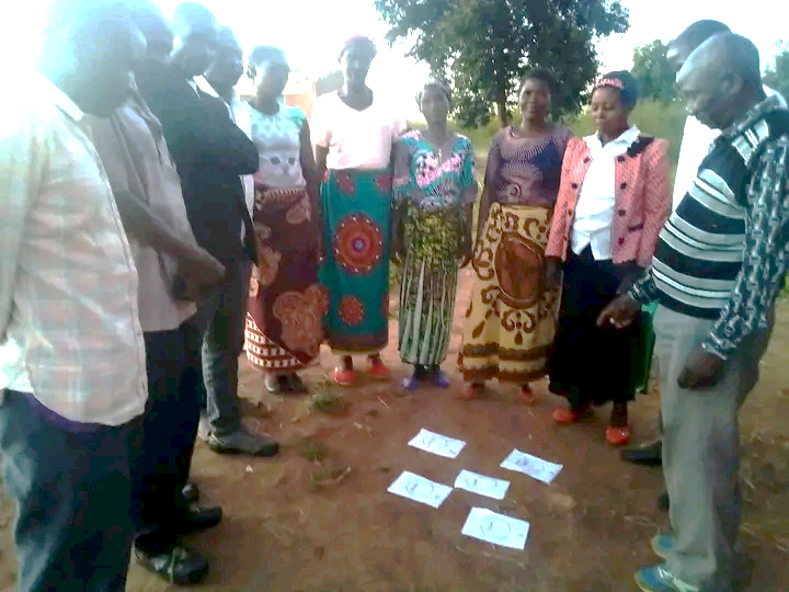 A group of person, standing in a half circle outside around several sheets of paper layed out on the ground.